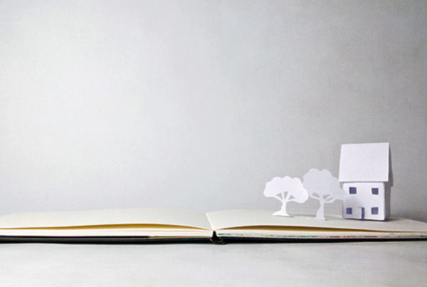 Tiny little hand-made paper house and two miniature trees on open book, sitting on white background.